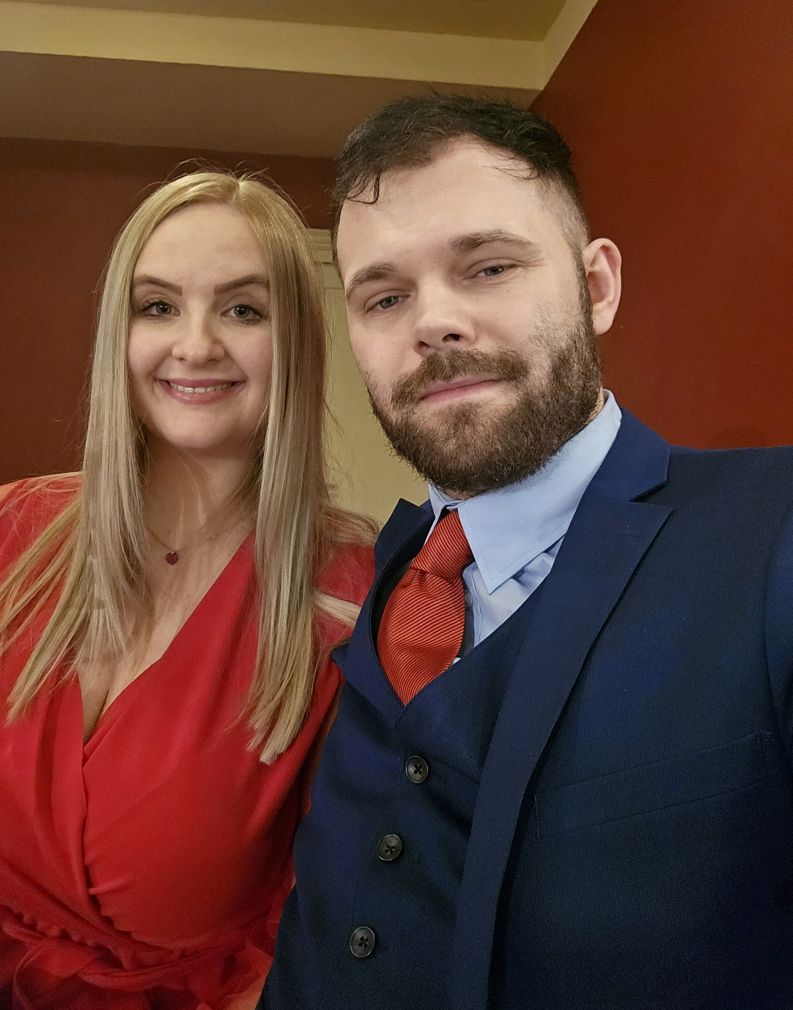 A smiling couple poses indoors. The woman wears a red dress, her hair long and blonde. The man wears a blue suit with a red tie, appearing content.
